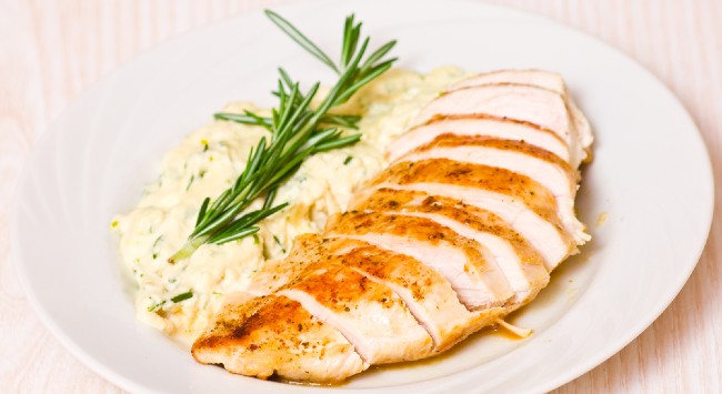 Filé de frango grelhado fatiado e servido com creme de milho com um ramo de lacrem em cima para decorar. Os elementos estão em um prato branco fundo.