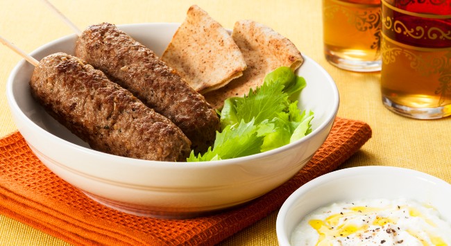 Espetinhos de kafta de carne em uma tigela branca, servida com pão sírio e alface. Na frente, uma outra tigela com molho de iogurte