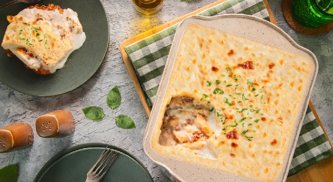 Assadeira com lasanha gratinada recheada com carne e molho branco. Ao lado, um prato verde com uma fatia da mesma lasanha.