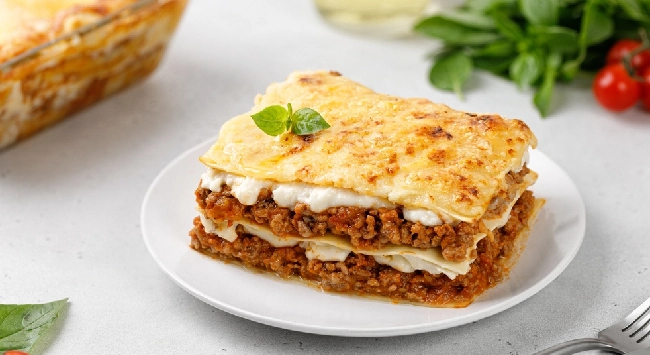 Fatia de lasanha de carne moída com molho branco de maizena em um prato redondo. Ao lado, uma travessa transparente com a mesma lasanha.