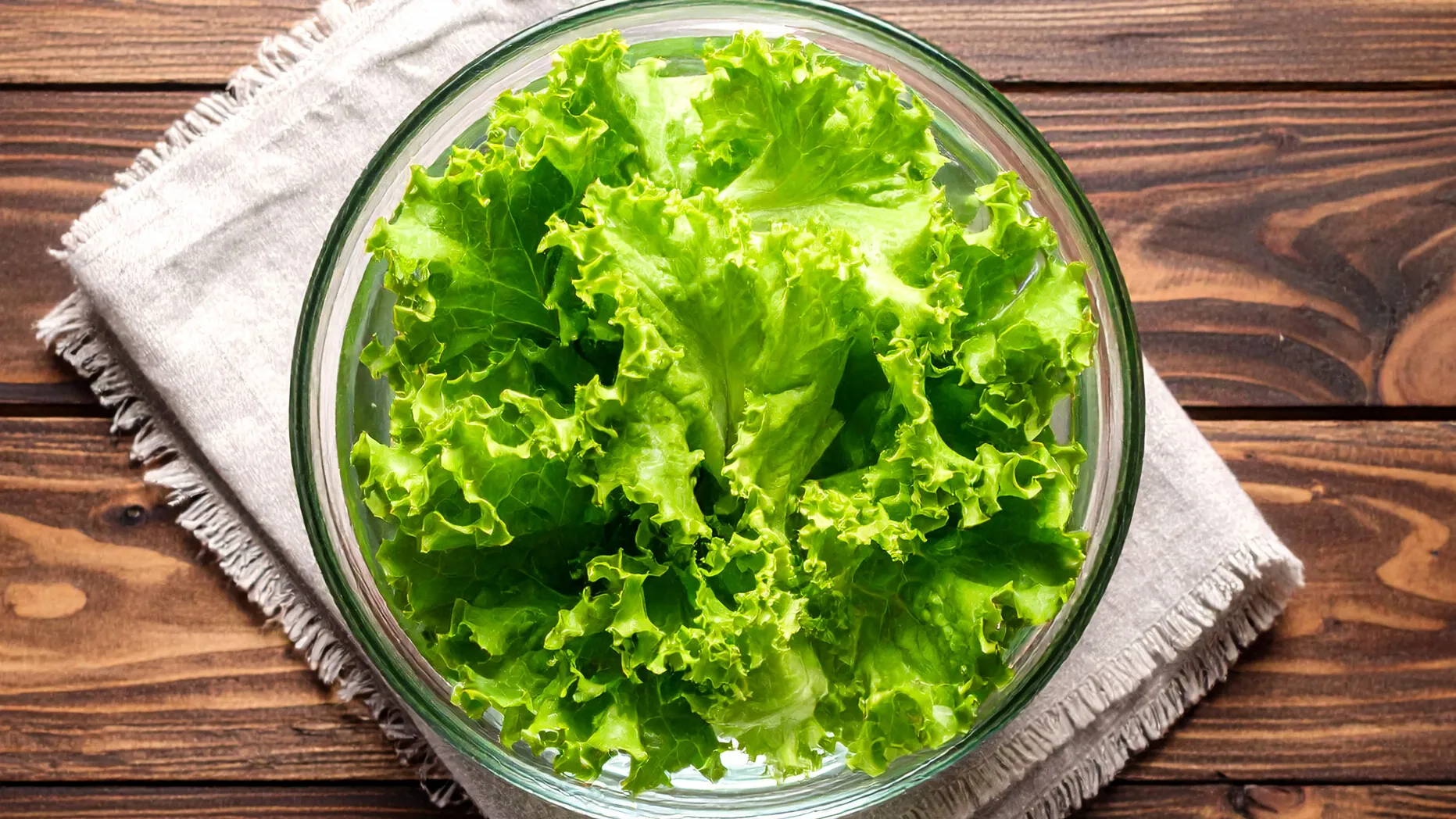 Folhas de alface verde fresca estão acondicionadas em uma tigela de vidro transparente, sobre um tecido de cor clara em uma superfície de madeira escura, ilustrando como conservar alface na geladeira.