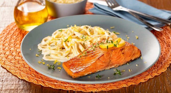 Filé de salmão com espaguete ao limone em um prato cinza. Ao fundo, um vidro de azeite.
