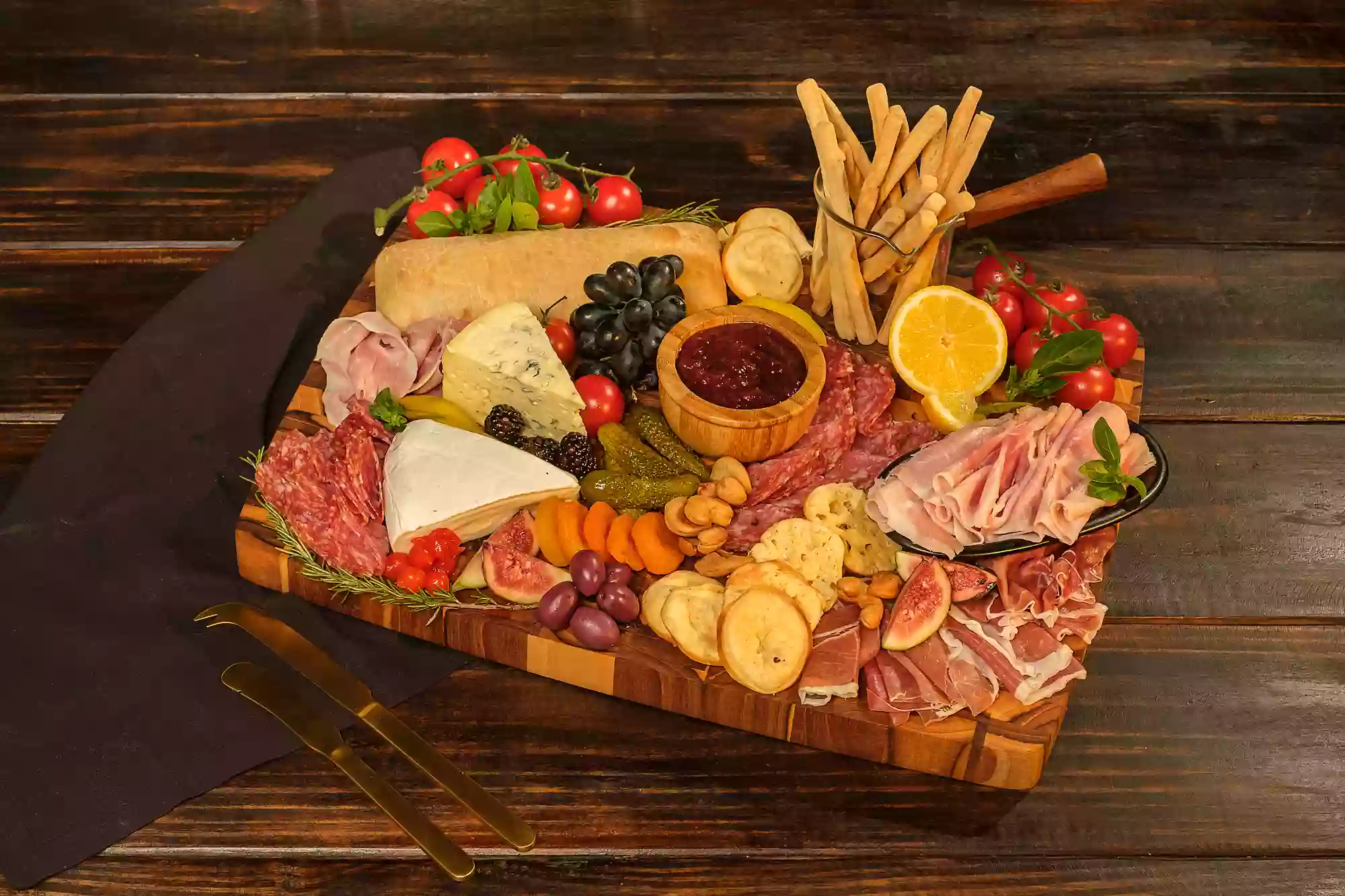 Imagem de uma tábua de frio com diversas entradas sob uma tábua de madeira quadrada, com um par de talheres ao lado, sob uma mesa de madeira