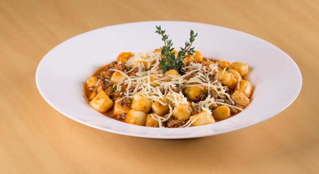 A imagem mostra um prato de nhoque à bolonhesa servido em um prato fundo branco. O nhoque, composto por pequenas massas de batata, está coberto com molho à bolonhesa de carne moída e tomate, além de ser finalizado com uma generosa porção de queijo ralado por cima. No centro do prato, há um ramo de ervas frescas, provavelmente tomilho, que serve como guarnição decorativa. A aparência do prato é apetitosamente rústica, com os cubos de nhoque bem destacados entre o molho e o queijo derretido, criando uma refeição convidativa e reconfortante.