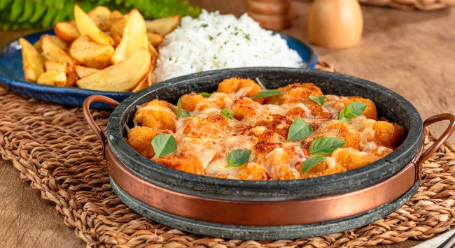 Parmegiana de camarão em uma panela de pedra. Ao fundo, um prato azul com arroz e batatas fritas.
