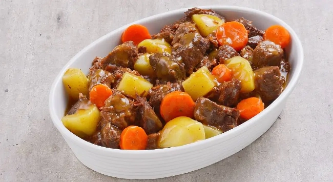 patinho servido em uma travessa oval branca. O ensopado é composto por pedaços generosos de carne bovina cozida, que parecem estar bem macios, acompanhados de pedaços de batata e rodelas de cenoura.
