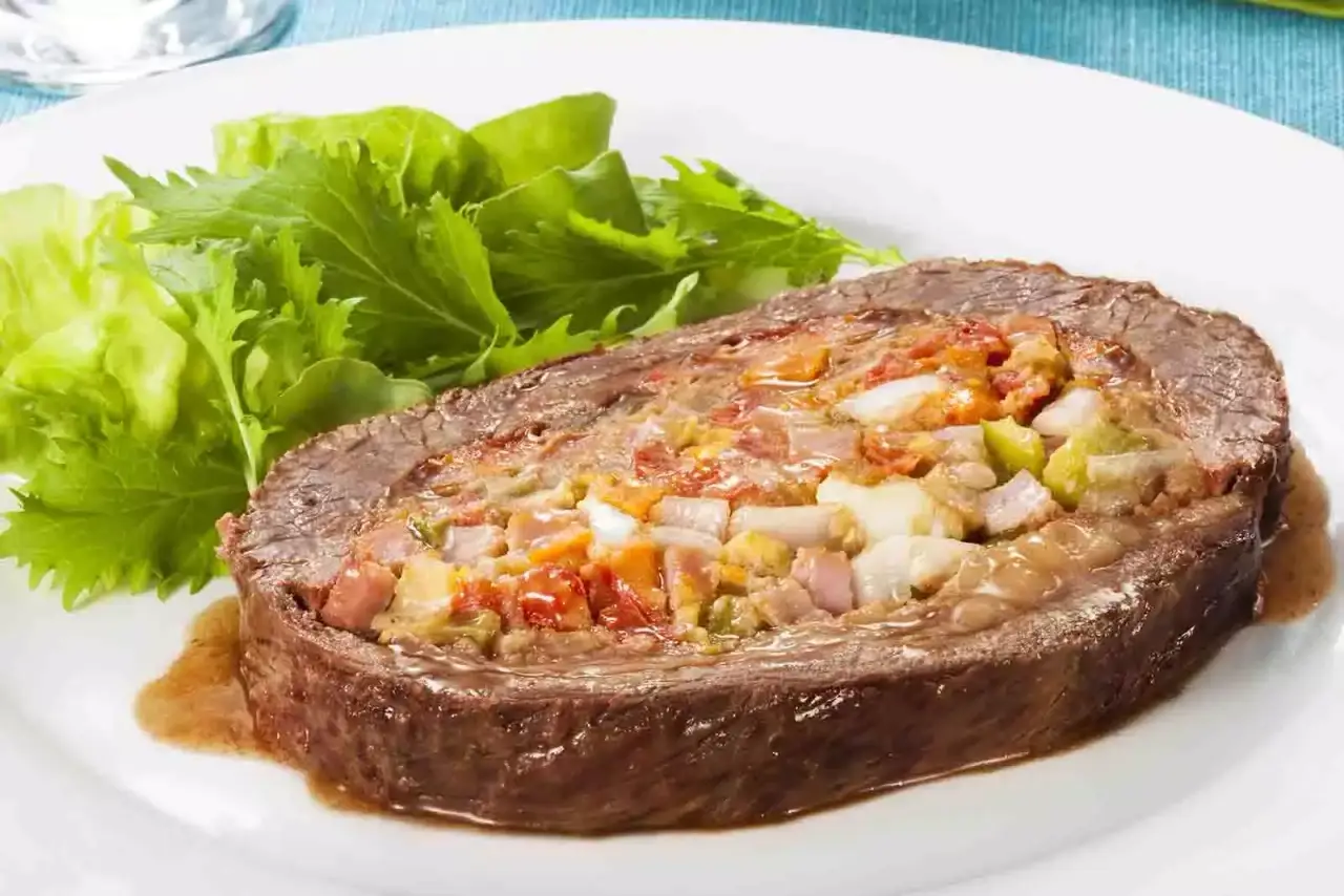 Imagem de uma fatia de picanha recheada sob um prato redondo branco, com folhas de salada ao lado, sob uma mesa azul