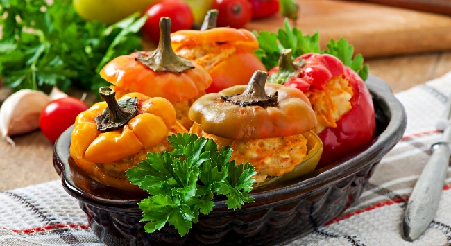 Travessa de pimentões recheados com frango. Ao fundo, ingredientes frescos como tomates e pimentões inteiros.