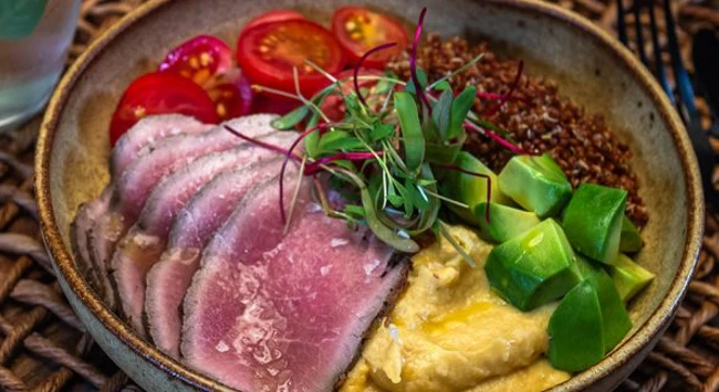Poke montado com rosbife, cubos de avocado, homus, tomatinhos e quinoa vermalha em uma tigela.
