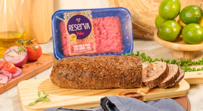 A imagem mostra um rocambole de carne moída assado, servido sobre uma tábua de madeira clara. O rocambole é marrom e levemente dourado, com uma superfície texturizada e temperada. Ao lado direito, algumas fatias do rocambole foram cortadas, revelando o recheio. Ao fundo, há uma embalagem azul com carne moída crua de uma marca chamada 'Reserva', parcialmente visível. Ao redor, ingredientes frescos como tomate, cebola roxa, e limões verdes estão dispostos em uma mesa de mármore, junto com uma garrafa de azeite, adicionando um toque colorido e natural à cena