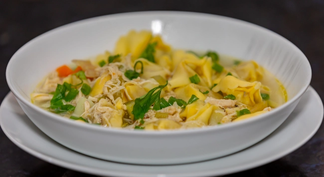 Sopa de agnoline servida em prato fundo branco, com caldo claro, frango desfiado, legumes em cubos e finalizada com cheiro-verde fresco.
