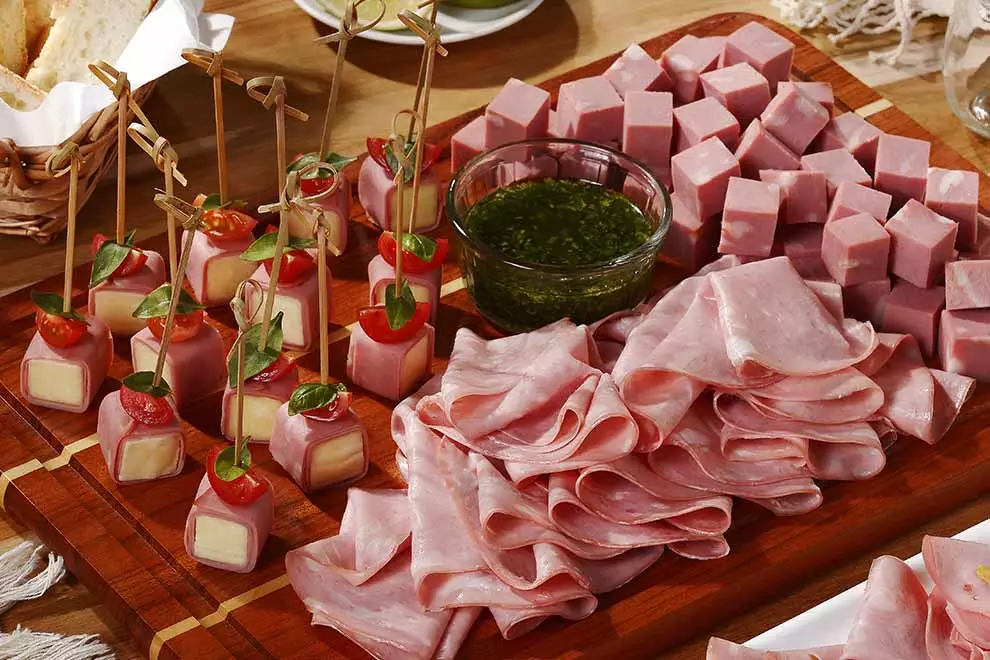 Tábua de frios com mortadela em formatos diferentes como em fatias, em cubos e enrolados em cubos de mussarela, sob uma tábua de madeira, com uma combuca de molho pesto no meio, em cima de uma mesa de madeira.