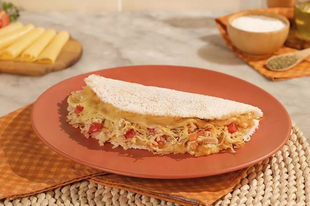 Imagem de uma tapioca de recheada com frango, sob um prato redondo, em cima de uma mesa de mármore.
