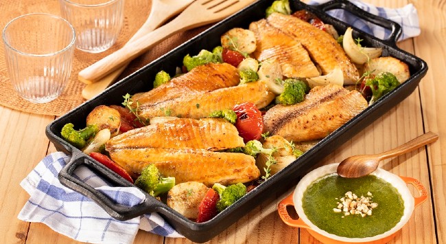 Travessa preta com filés de tilápias e uma salada diversa de legumes com molho pesto para acompanhar.