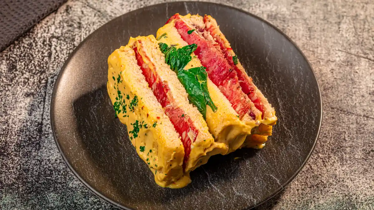 Uma torta fria com camadas de pão, salame, creme amarelo e folhas verdes, servida em um prato escuro sobre uma superfície texturizada.