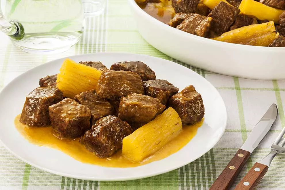 Imagem de um prato redondo branco com vaca atolada, com um par de talheres ao lado e uma travasse branca com vaca atolada ao fundo, sob uma mesa com toalha branca com listras verdes